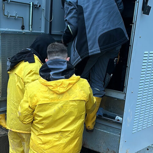 Personen in gelben Regenjacken arbeiten an einem Gerät im Freien, umgeben von grauer Technik.