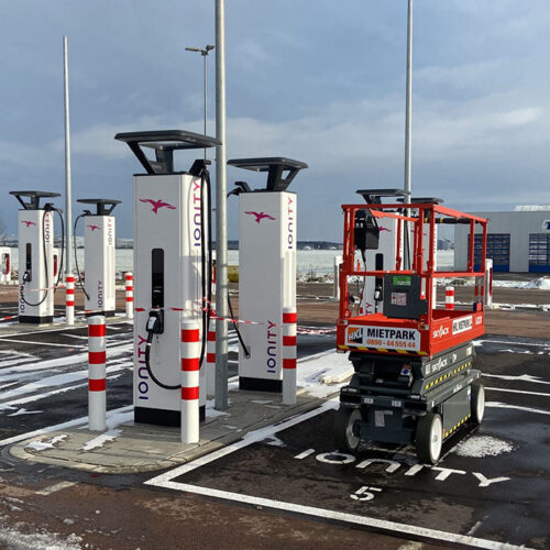 Ladestationen auf leerem Parkplatz, Hebebühne rechts, bewölkter Himmel, einige Schneeflecken.