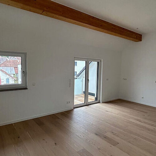 Großer, leerer Raum mit Holzfußboden, weißen Wänden, Holzträgerdecke und Zugang zu einem Balkon.