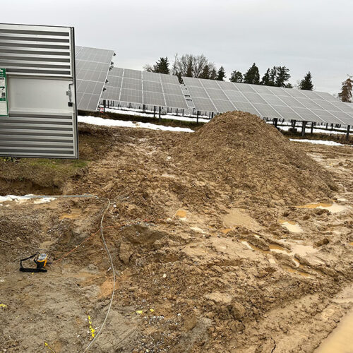 Solarfeld bei bewölktem Himmel, Matsch im Vordergrund, geöffneter Schaltkasten links sichtbar.