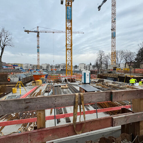 Baustelle mit Kränen, Bauarbeitern in gelben Westen und winterlichen Bäumen im Hintergrund.