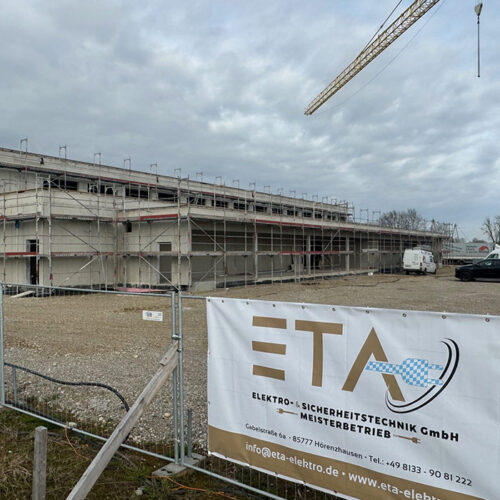 Eine Baustelle mit grauem Gebäude, Gerüst, gelbem Kran und Firmenbanner im Vordergrund.