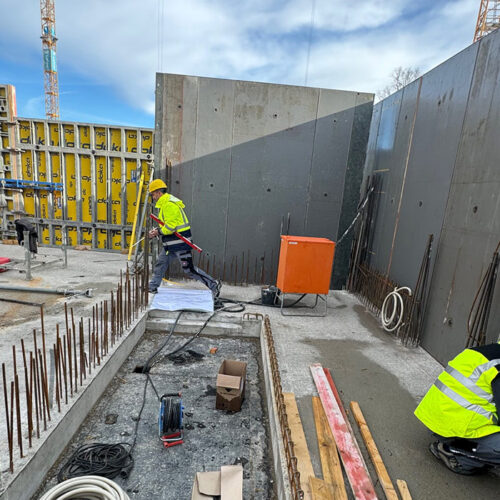 Drei Bauarbeiter auf einer Baustelle, tragen helle Schutzwesten, arbeiten an Betonwänden.