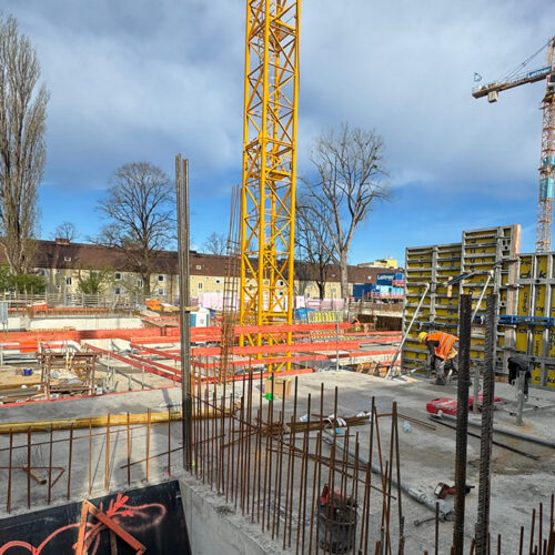 Baustelle mit Kran, Arbeiter in orange, strahlend blauer Himmel, viele Stahlträger in einem Gebäude.