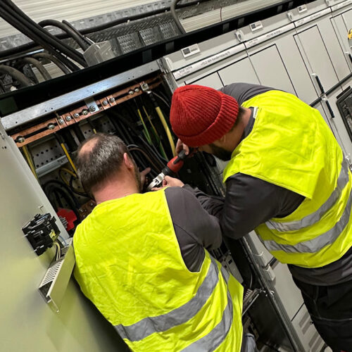 Zwei Personen in gelben Westen arbeiten konzentriert an Elektrokabeln in einem Technikraum.