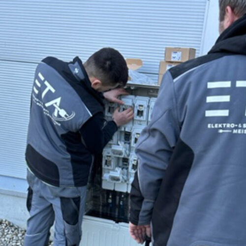 Zwei Handwerker in grauen Overalls arbeiten an einem Sicherungskasten mit konzentriertem Ausdruck.