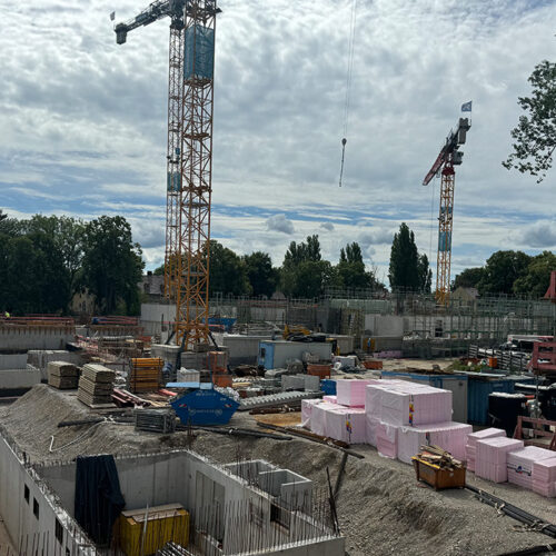 Baustelle mit Kränen und Baumaterialien, bewölkter Himmel, Gebäude im Hintergrund zu sehen.