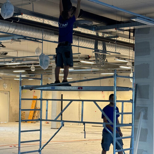 Eine Person auf Baugerüst, arbeitet an der Decke; andere steht daneben in leerem Raum.