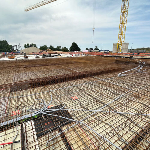 Baustelle mit Stahlgitter und Baukran, trüber Himmel, einzelne Arbeiter in der Ferne.