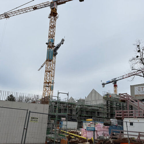 Baustelle mit Kränen, Gerüsten und Bauzaun, bedeckter Himmel. Keine Personen sichtbar.