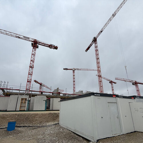 Baugebiet mit mehreren roten Kränen und weißen Containern unter grauem Himmel.