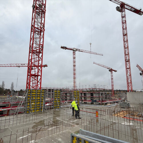 Eine Baustelle mit mehreren roten Kränen; ein Arbeiter in gelber Weste steht auf dem Betonboden.