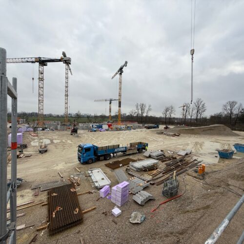 Eine Baustelle mit Kränen, LKW und Baumaterialien unter bewölktem Himmel.