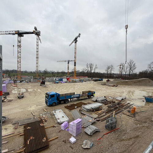 Baustelle mit zwei Kränen, Lastwagen und Baumaterialien, bewölkter Himmel.