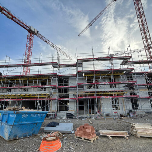 Baustelle mit Kranen und Gerüst. Mehrstöckiger Rohbau unter blauem Himmel, Baumaterial vorne.