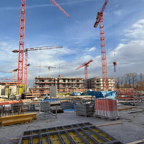 Baustelle mit roten Kränen vor blauem Himmel, Baugerüste und Baumaterialien verstreut.