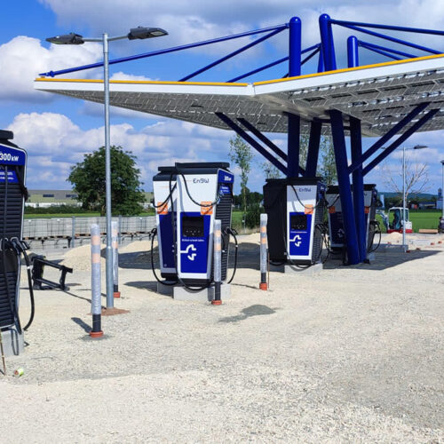 Eine moderne blaue E-Ladestation mit drei Ladepunkten unter einem Solardach bei sonnigem Wetter.