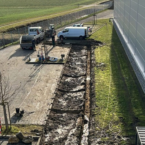 Bauarbeiter an Grube neben grauem Gebäude, umgeben von geparkten Fahrzeugen und offener Landschaft.